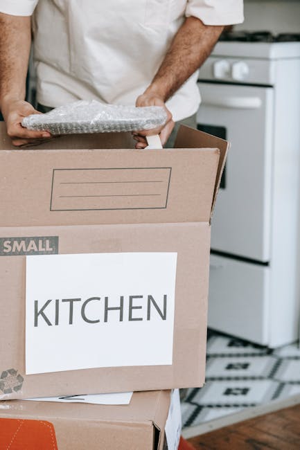 A person in a white shirt is seen loading a cardboard box labeled 'KITCHEN' onto a moving van or cart inside a home. The box is partially open, revealing bubble wrap used for packing fragile items. Behind the person, part of a white kitchen stove and oven are visible, indicating the move involves household belongings. The setting appears to be a kitchen area with patterned floor tiles, and the person is carefully carrying the box, suggesting a home relocation or furniture transport process. This scene illustrates packing and loading activities associated with professional removals services provided by Man With a Van Plumstead, especially for house moves involving furniture transport and packing logistics.