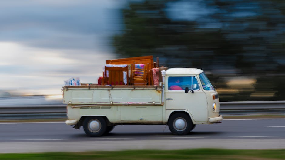 A vintage white delivery van is in motion on a road during daylight, with a blurred background indicating speed. The van's cargo area is loaded with various household items, including a wooden dining chair, a small wooden table, and several cardboard boxes wrapped in plastic. Other packing materials such as fabric covers and protective blankets are also visible, securing the belongings for transport. The scene captures the loading process involved in home relocation or furniture transport, with the van parked near a curb and the items carefully arranged to maximise space. Man With a Van Plumstead specialises in removals and professional moving services, supporting clients with packing, loading, and transporting belongings efficiently and safely, as shown by the careful stacking of furniture and boxes in this moving scenario.