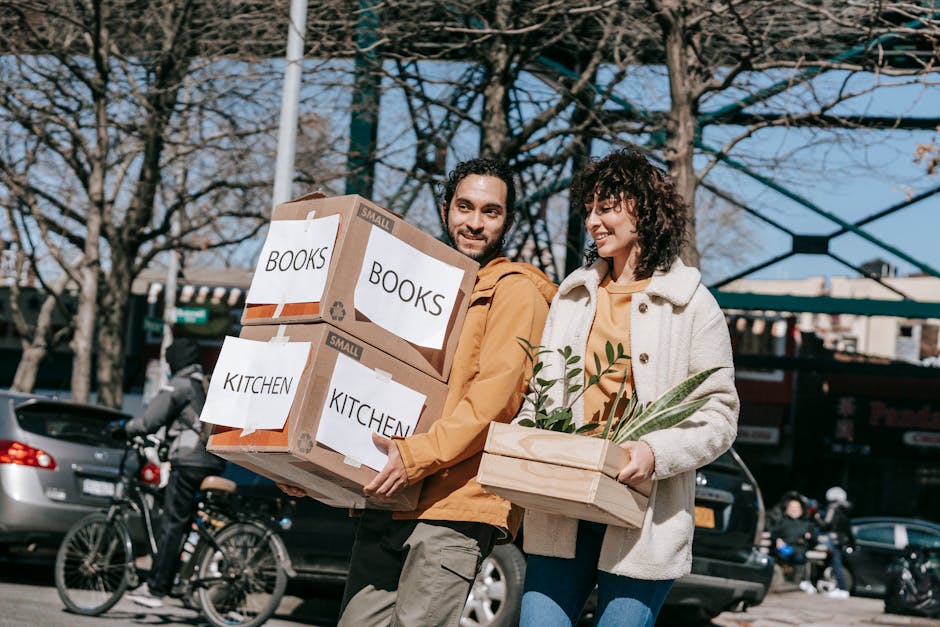 A person in a white shirt is seen loading a cardboard box labeled 'KITCHEN' onto a moving van or cart inside a home. The box is partially open, revealing bubble wrap used for packing fragile items. Behind the person, part of a white kitchen stove and oven are visible, indicating the move involves household belongings. The setting appears to be a kitchen area with patterned floor tiles, and the person is carefully carrying the box, suggesting a home relocation or furniture transport process. This scene illustrates packing and loading activities associated with professional removals services provided by Man With a Van Plumstead, especially for house moves involving furniture transport and packing logistics.