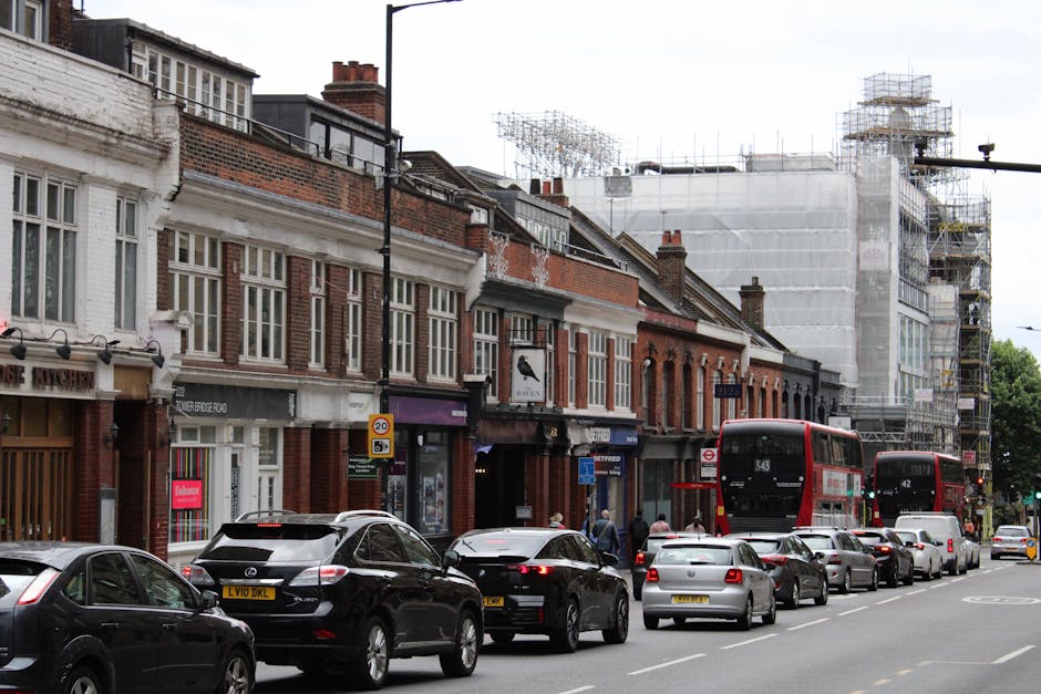 A busy street scene on Plumstead High Street featuring a row of brick-fronted shops with large display windows and signage, some with awnings, lining the sidewalk. Several vehicles, including black and white cars and two red double-decker buses, are parked or moving along the street. In the foreground, a line of parked cars extends along the curb, while pedestrians are visible walking on the pavement. Behind the shops, a building under construction is covered in white scaffolding and netting, with multiple levels and construction equipment. The street appears to be part of a home relocation or furniture transport process, with the scene capturing the loading and moving environment around Man With a Van Plumstead, as it prepares for or during a house removals service on Plumstead High Street.