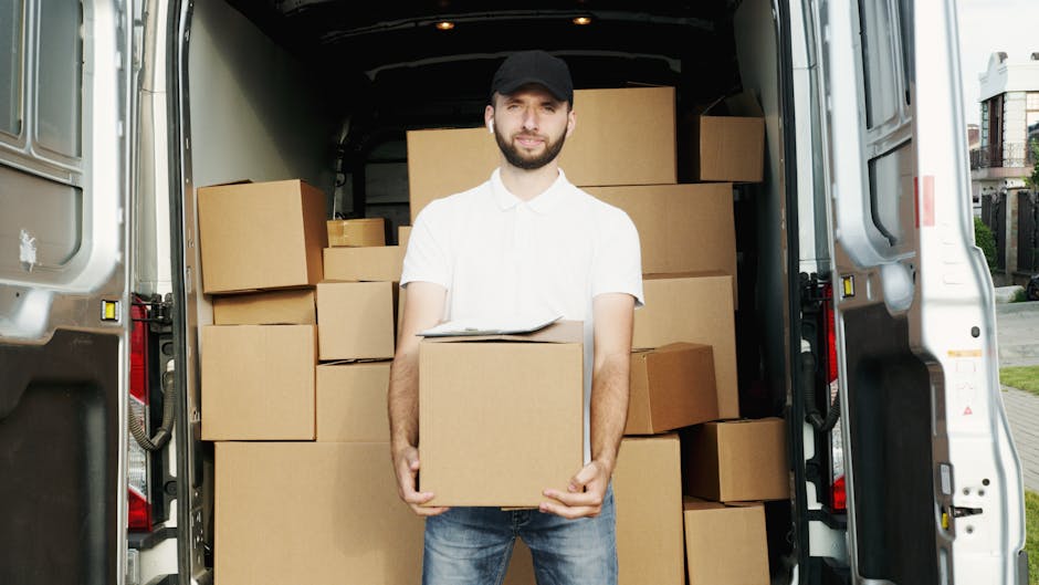 A man with a beard, dressed in a white shirt, jeans, and a black cap, is standing inside the open rear of a white moving van, holding a medium-sized cardboard box. The van is filled with several similar cardboard boxes of various sizes, some stacked on top of each other, used for packing and transporting household belongings during a home relocation. The containers are secured with plastic wrap and are positioned on the van's floor and walls, indicating an active loading process. Outside the van, the pavement and a residential area with houses and greenery are visible, suggesting the moving activity is taking place in a residential neighbourhood. The lighting is natural, providing clear visibility of the boxes, the man, and the surrounding environment. This image reflects professional furniture transport and packing services offered by Man With a Van Plumstead, focused on efficient removals within the SE18 area.