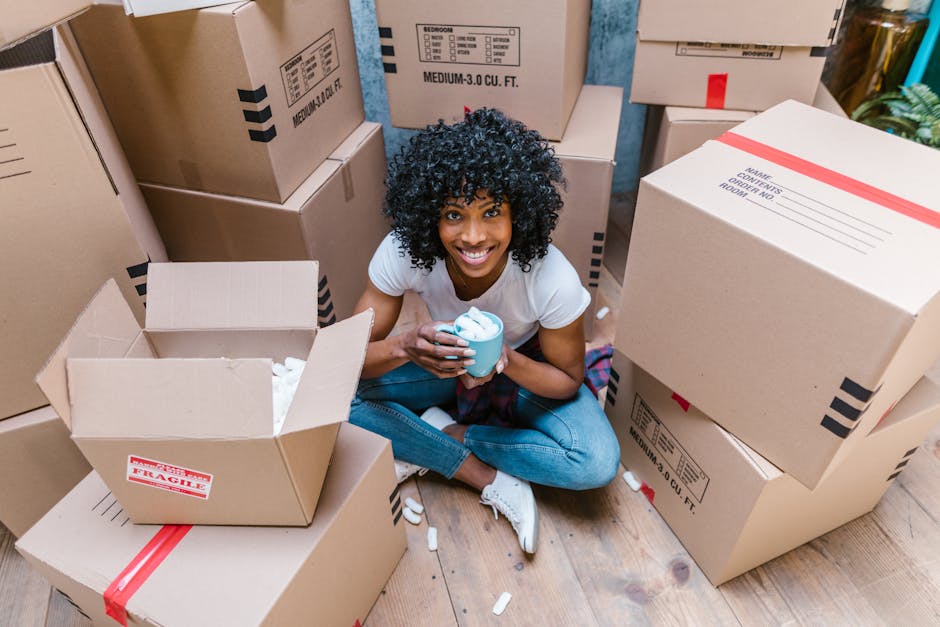 A young woman with curly black hair, wearing a white t-shirt, blue jeans, and white sneakers, is sitting on a wooden floor surrounded by several packed cardboard boxes used for house removals. She is smiling and holding a blue mug, with packing materials such as crumpled paper and foam inserts scattered around. The boxes display labels for contents and fragile warnings, and some are open showing tissue paper wrapping. The scene is indoors, likely within a home or a storage area, with ambient natural light. The image depicts the packing stage of a home relocation, a process often managed by professional removal services like Man With a Van Plumstead, focusing on careful packing and organising before furniture transport and moving day.