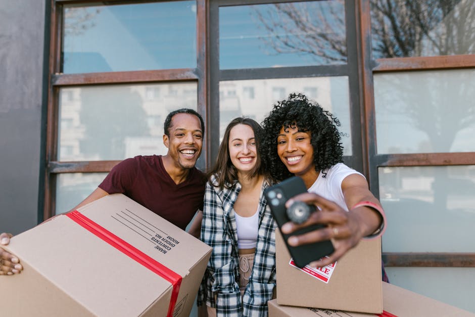 A group of three smiling individuals standing outdoors in front of a modern building with large windows, holding cardboard boxes sealed with red tape, likely containing household items for moving. The person on the left, a man wearing a maroon t-shirt, holds a box labeled 'wide dimensions,' while the woman in the middle, with long brown hair, is dressed in a plaid shirt and white top. The woman on the right, with curly black hair, is wearing a white t-shirt with rolled-up sleeves and is taking a selfie with a smartphone, capturing the group and boxes. They are positioned on a paved surface with some greenery visible in the background. This scene reflects the packing and home relocation process, part of a moving or removals service provided by Man With a Van Plumstead, emphasizing the preparation stage of furniture transport and logistics for house removals.