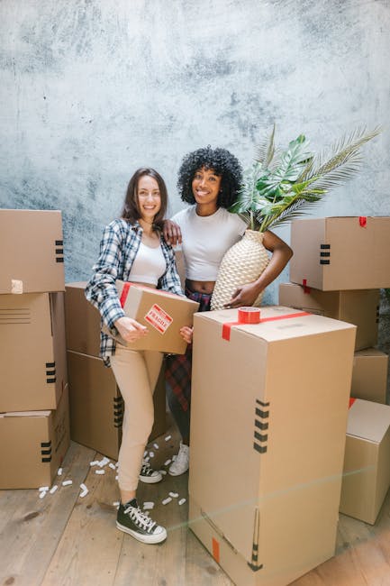 A young woman with curly black hair, wearing a white t-shirt, blue jeans, and white sneakers, is sitting on a wooden floor surrounded by several packed cardboard boxes used for house removals. She is smiling and holding a blue mug, with packing materials such as crumpled paper and foam inserts scattered around. The boxes display labels for contents and fragile warnings, and some are open showing tissue paper wrapping. The scene is indoors, likely within a home or a storage area, with ambient natural light. The image depicts the packing stage of a home relocation, a process often managed by professional removal services like Man With a Van Plumstead, focusing on careful packing and organising before furniture transport and moving day.
