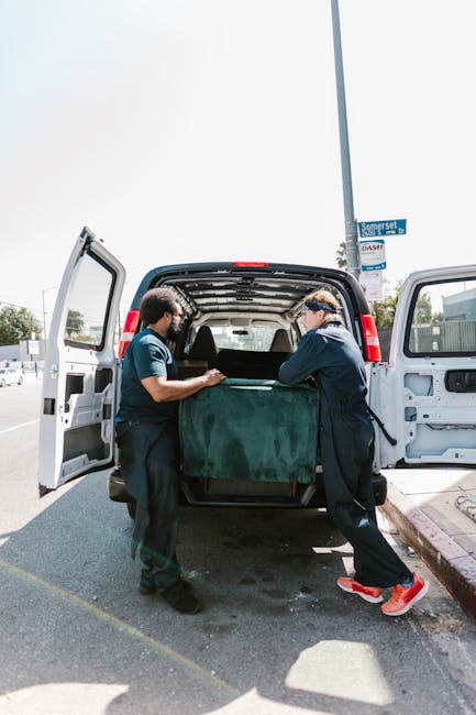 Two movers from Man With a Van Plumstead are engaged in the loading process of a home relocation, carefully lifting and positioning a large, green upholstered furniture item into the rear cargo area o