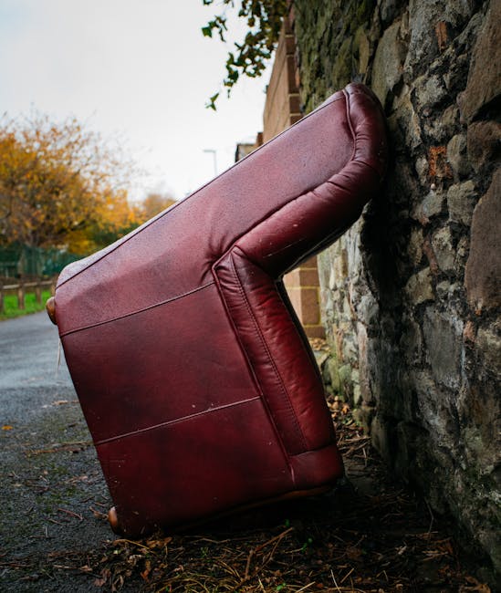 A red fabric sofa with visible wear and crease marks rests against a stone wall outdoors on a pavement. The sofa appears to be mid-removal, positioned at an angle with its back against the wall and the front facing outward. Surrounding the sofa are scattered leaves and dirt, indicating an outdoor setting. In the background, there is a wooden fence and trees with orange and green foliage, suggesting an autumn day. The scene is illuminated by natural light, with a partly cloudy sky visible in the distance. This image captures a furniture removal process, possibly part of a home relocation or disposal service by Man With a Van Plumstead, illustrating the steps of loading household furniture for transport, such as sofa disposal or moving tasks involved in domestic removals.