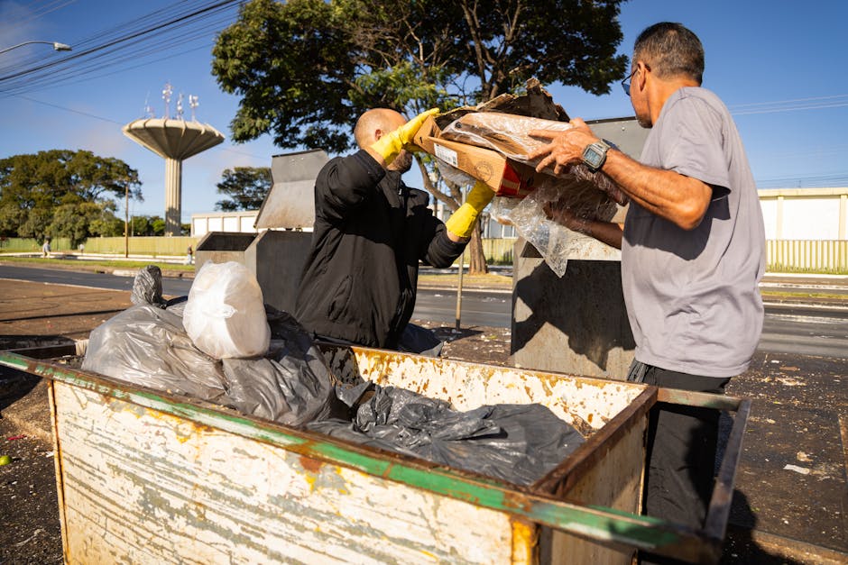 Two men are engaged in the disposal process of an old sofa as part of home relocation or clearance in SE18, Plumstead. They are positioned next to a large metal waste container located outdoors on a paved street, with one man pouring materials from a cardboard box wrapped in plastic, while the other man, wearing a watch and gloves, assists in the loading. Inside the waste container, there are black and grey garbage bags and other debris related to furniture removal. Behind them, a backdrop of trees, a water tower, and a white fence are visible, indicating a residential or mixed-use area. The men are dressed in casual clothing suitable for manual work, and the scene is illuminated by bright daylight. This image demonstrates the process of loading bulky furniture like sofas for disposal or transfer during a furniture transport or moving service, with attention to proper handling and material containment, aligned with the services provided by Man With a Van Plumstead.