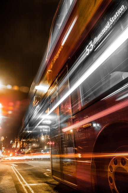 Nighttime scene of a double-decker bus in motion, with light trails indicating speed and movement. The bus displays the route number 53 and destination 'Plumstead Station' on the front, with the route information illuminated against the dark background. Streetlights and passing vehicle headlights create additional light trails on the road, which is marked with double yellow lines along the curb. The image captures the dynamic atmosphere of urban transportation, relevant to house removals and relocation services provided by Man With a Van Plumstead, illustrating the logistical aspects of moving and transportation within the area.