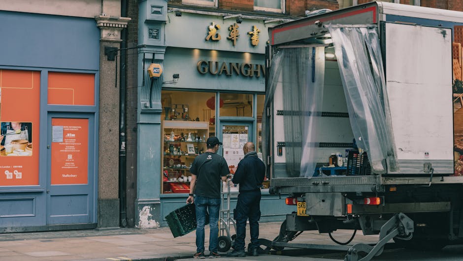 Two men are engaged in the loading process outside a commercial building with signage indicating a store named 'GUANGH'. One man is holding a black crate, while the other appears to be conveying a small trolley or dolly. A large delivery van is parked nearby, with its side door open, revealing a partially covered cargo area with transparent plastic sheeting. The van is positioned on a paved sidewalk in front of the store, which has large glass windows and a blue exterior finish. The building’s facade features a decorative cornice and stone detailing around the entrance, with additional signage displaying Chinese characters above the store window. Inside, the store's interior is visible through the glass, showing shelves with various products. The scene captures the typical logistics involved in house or business relocations, highlighting furniture transport and packing activities central to professional removals services, with Man With a Van Plumstead occasionally performing such tasks.