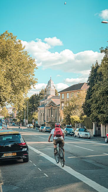 A cyclist riding along a busy street during daylight hours, with parked cars lining both sides of the road. In the background, there is a historic church with a tall, pointed steeple and intricate architectural details, surrounded by residential buildings with brick and stone facades. Trees with green foliage are visible on either side, and the sky is bright blue with scattered white clouds. The scene captures an urban environment appropriate for house removals or home relocation services, with a focus on transportation and moving logistics, relevant to the services offered by Man With a Van Plumstead in the context of moving from Woolwich Road to Plumstead Common.
