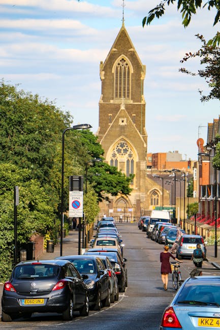 A row of parked cars lines both sides of a residential street with a church featuring a tall, pointed steeple and large arched windows in the background. The street is lined with trees and lampposts, with one person walking with a bicycle and another carrying a bag. The scene is set during daylight under a partly cloudy sky. This image illustrates the typical environment for home relocation or moving logistics in Plumstead, captured near Woolwich Road. Man With a Van Plumstead’s moving services often involve loading furniture, boxes, and appliances onto a van parked nearby, with the process including careful packing in cardboard boxes, wrapping furniture in blankets or plastic wrap, and using trolleys to transfer items from the property to the vehicle. The street's calm, suburban atmosphere reflects the setting where professional removals take place, supporting efficient furniture transport and home relocation activities within the area.