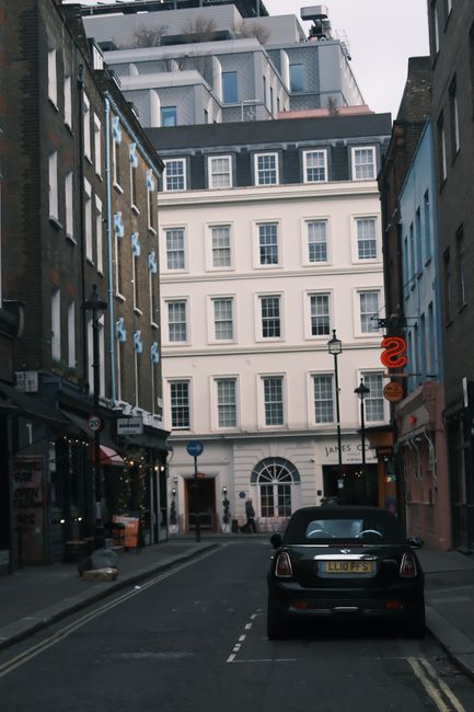 A narrow street lined with multi-storey buildings, including a white three-story structure with numerous windows and an arched entrance at the ground level, seen from a ground-level perspective. The street features parked vehicles, with a black van positioned on the right side in the foreground, indicating active loading or unloading. On the street, there are a few small objects such as bags or boxes, and street lamps are visible attached to the buildings. The area appears to be in an urban environment with a mixture of classic brick facades and modern rooftop extensions visible at the top of the image. This setting reflects an ongoing home relocation or furniture transport process, showcasing the typical environment in which house removals and moving services like those provided by Man With a Van Plumstead operate, particularly during packing, loading, or transport stages involving boxes, packaging materials, and furniture being moved through the lane.
