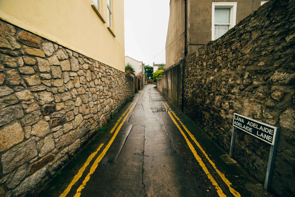 A narrow residential alleyway in Plumstead with a wet, dark asphalt surface, flanked by stone and brick walls on each side. The alley features double yellow lines painted along the edges, indicating parking restrictions. At the foreground, a small sign displaying the street name 'LANA ADELAIDE' in both English and Welsh leans against the stone wall on the right. The alley extends into the distance, with some greenery and residential buildings visible at the far end, under an overcast sky. The scene suggests an urban environment suitable for home relocation or furniture transport, with enough space for moving vehicles and careful handling of packed boxes or household furniture during a house removal process, as managed by companies like Man With a Van Plumstead.
