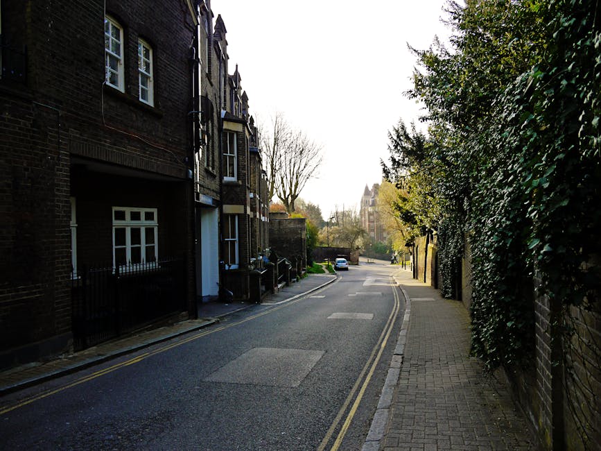 A narrow street lined with multi-storey buildings, including a white three-story structure with numerous windows and an arched entrance at the ground level, seen from a ground-level perspective. The street features parked vehicles, with a black van positioned on the right side in the foreground, indicating active loading or unloading. On the street, there are a few small objects such as bags or boxes, and street lamps are visible attached to the buildings. The area appears to be in an urban environment with a mixture of classic brick facades and modern rooftop extensions visible at the top of the image. This setting reflects an ongoing home relocation or furniture transport process, showcasing the typical environment in which house removals and moving services like those provided by Man With a Van Plumstead operate, particularly during packing, loading, or transport stages involving boxes, packaging materials, and furniture being moved through the lane.