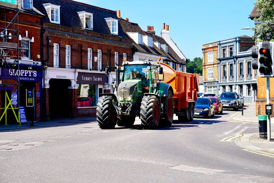 A large green tractor with oversized tires is positioned at an urban intersection in a residential area, attached to an orange waste collection container on its rear. The tractor is part of a home relocation or waste collection process, with the loading area next to a sidewalk lined with brick and stucco buildings. The scene is well-lit with bright sunlight, casting minimal shadows, and shows several parked cars along the street. The background includes a traffic light on a pole, shopfronts with signs such as 'Sloppy's Sports Bar' and 'Tierney Salons,' and trees providing greenery. This setup suggests the tractor is engaged in moving heavy loads, possibly related to local services like removals or waste clearance, with the surroundings indicating a typical town or city street in Plumstead, where proper council permit rules for large vehicles such as these are necessary.