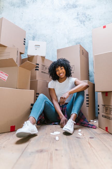 A young woman with long brown hair, smiling and wearing a plaid shirt and beige trousers, stands indoors against a textured light blue wall during a home relocation process. She holds an empty medium-