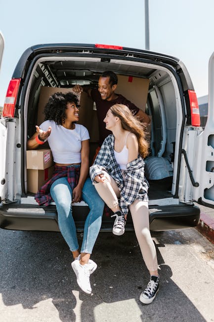 Two professional movers from Man With a Van Plumstead are loading a large, green upholstered piece of furniture into the cargo area of a white van, which is parked on a street with a sidewalk and stre