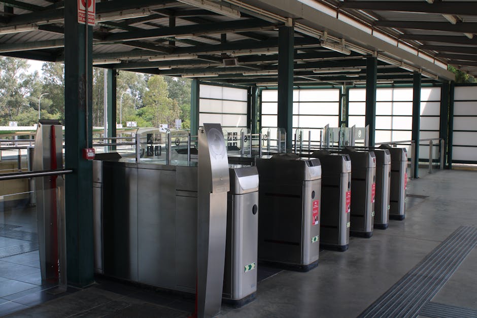 Interior view of a parking area at Plumstead Station featuring a row of stainless steel ticket barriers with black arms and red warning labels, positioned along a smooth concrete floor. The barriers are arranged in alignment, with each barrier equipped with a card reader or access control panel visible on the front. Surrounding the area are metallic and glass panels forming safety railings, supported by green metal columns that extend to the ceiling, which consists of exposed beams and structural elements. Large windows and semi-opaque panels create natural lighting conditions, illuminating the space. Outside the station, visible through the windows, are trees and a traffic road, indicating an outdoor setting. The scene captures a clean, orderly environment commonly encountered during station relocations or preparations for moving equipment, which Professional removals services like Man With a Van Plumstead may assist with during furniture transport or station access logistics.