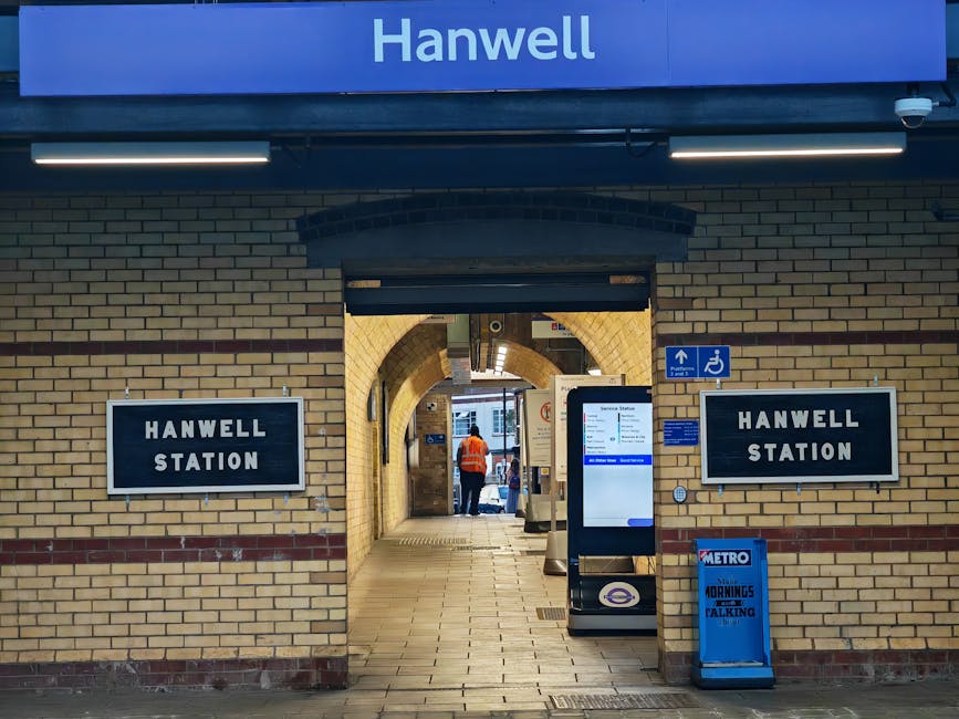 Interior view of a parking area at Plumstead Station featuring a row of stainless steel ticket barriers with black arms and red warning labels, positioned along a smooth concrete floor. The barriers are arranged in alignment, with each barrier equipped with a card reader or access control panel visible on the front. Surrounding the area are metallic and glass panels forming safety railings, supported by green metal columns that extend to the ceiling, which consists of exposed beams and structural elements. Large windows and semi-opaque panels create natural lighting conditions, illuminating the space. Outside the station, visible through the windows, are trees and a traffic road, indicating an outdoor setting. The scene captures a clean, orderly environment commonly encountered during station relocations or preparations for moving equipment, which Professional removals services like Man With a Van Plumstead may assist with during furniture transport or station access logistics.