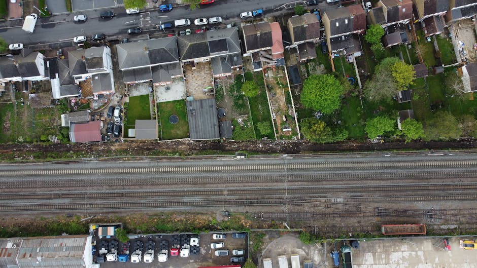 An aerial view of a residential area showing a row of houses with backyards, some with outdoor furniture, decking, and small garden features. A few trees are visible among the properties, with the houses situated directly adjacent to each other. Running parallel to the row of houses are railway tracks, separated by a narrow strip of land, with a few parked vehicles and an industrial building at the bottom edge of the image. The lighting appears natural, indicating daytime. This scene relates to house removals and moving logistics, with the railway and surrounding environment providing context for transport and clearance considerations often involved in home relocation services offered by Man With a Van Plumstead.
