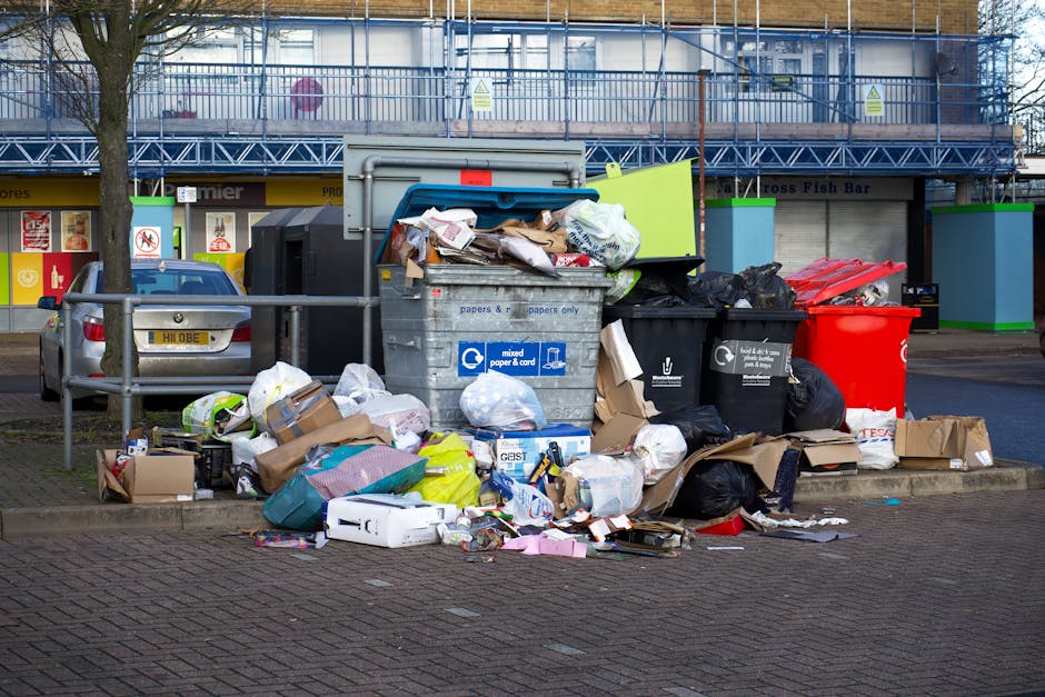 A collection of overflowing public waste bins and scattered rubbish, including cardboard boxes, plastic bags, paper packaging, and discarded items, positioned on a paved area near a metal railing on the street in Plumstead. Behind the bins, there are parked cars, and in the background, a building façade with scaffolding and a storefront with signage. The scene is illuminated by natural daylight, reflecting typical urban street conditions associated with waste management during house removals or relocation activities, as seen in professional moving services by Man With a Van Plumstead.