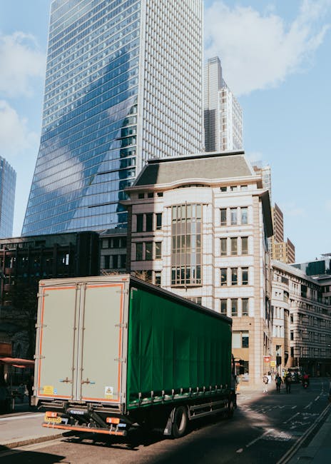 A large green delivery truck is parked on a city street in Plumstead, surrounded by tall modern glass office buildings and older, traditional multi-storey structures with detailed facades. The truck's rear doors are closed, with no visible loading activity, while the vehicle is positioned on the pavement near the curb. The scene is illuminated by daylight, with clear blue skies overhead, indicating a bright and mild day. This environment is typical of urban home relocation or furniture transport logistics, where Man With a Van Plumstead offers removals services, including vehicle loading, handling boxes, and transporting household items through busy city streets. The image captures the essence of professional moving processes in a city environment, highlighting the importance of careful loading, timing, and efficient vehicle use during home removals.