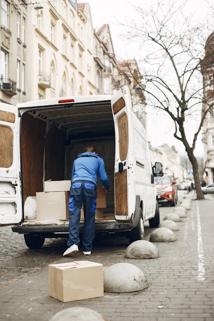 A man wearing a blue jacket and navy trousers is standing inside the open rear cargo area of a white moving van parked on a street. The van's interior wall panels are visible, and he appears to be arranging or loading large, wrapped items, possibly furniture or boxes, with some materials secured with plastic wraps. On the pavement nearby, a cardboard box rests on the ground, while the curb is lined with round concrete bollards. In the background, there are parked cars along the street, multi-storey residential buildings with ornate facades, and leafless trees, indicating a location in a city area. The scene is captured during daylight with overcast skies, and the environment suggests a home relocation or furniture transport process involving packing and loading. The image depicts a professional moving operation, reminiscent of services provided by companies like Man With a Van Plumstead, emphasizing the logistical aspects of house removals and moving services.